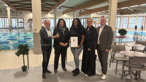 Fünf Personen stehen nebeneinander in einem Hallenbad. Die mittlere Person hält einen Förderbescheid in den Händen.