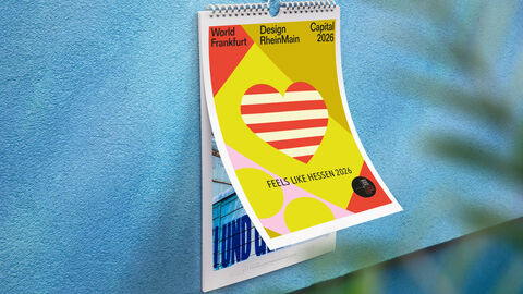 Ein Kalender hängt an einer weißen Hauswand. Das Deckblatt ist gelb mit einem roten Herz. Darauf steht geschrieben "Feels like Hessen 2026".