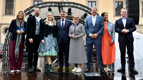 Eine Gruppe von acht festlich gekleideten Personen steht lächelnd nebeneinander auf einer nassen Open-Air-Bühne. Unter ihnen befindet sich eine Frau in einem auffälligen, blau-grünen Trachtenkleid mit Blumenkranz. Im Hintergrund ist ein historisches Schlossgebäude zu sehen.
