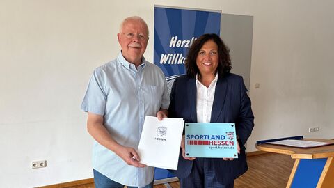 Karin Müller steht neben einem älteren Herrn, er hält einen Förderbescheid und sie ein Schild mit der Aufschrift "Sportland Hessen" in den Händen.