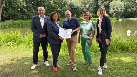 Fünf Personen stehen nebeneinander an einem See-Ufer. Zwei halten gemeinsam einen Förderbescheid in den Händen.