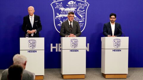 Herr Schwarz, Herr Rhein und Herr Mansoori stehen jeweils an einem eigenen Rednerpult - im Hintergrund das Wappen von Hessen