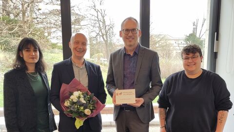 Sozialminister Kai Klose und Knud Wechterstein stehen mit zwei weiteren Personen vor Fenster. Kai Klose hält Urkunde in der Hand.