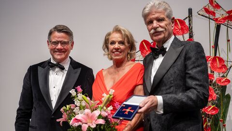 Ministerpräsident Boris Rhein und Sonja und Reinhard Ernst schauen in Kamera. Reinhard Ernst trägt das Bundesverdienstkreuz, Sonja Ernst einen Blumenstrauß