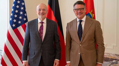 Zwei Männer stehen vor Flaggen der USA, Deutschland und Hessen.