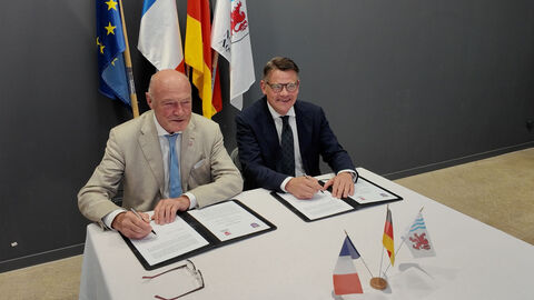 Zwei Männer sitzen nebeneinander an einem Tisch und sind gerade dabei ein Dokument zu unterzeichnen.