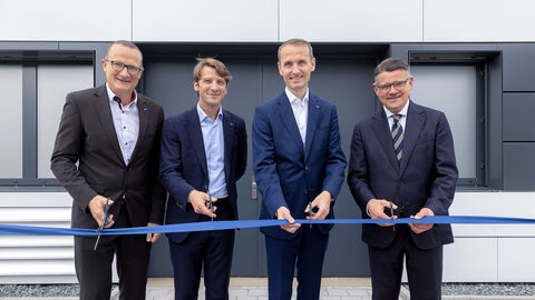 Vier Männer stehen in Anzügen gekleidet nebeneinander. Vor ihnen ist ein blaues Band gespannt, das sie mit einer Schere durchschneiden wollen. 