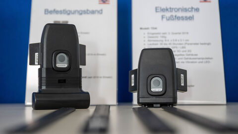 Zwei elektronische Fußfesseln liegen vor erklärenden Aufstellern.