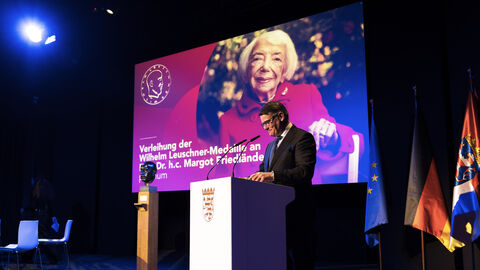 Person steht an Rednerpult. Auf dem Bildschirm im Hintergrund sieht man ein Foto einer älteren Frau.